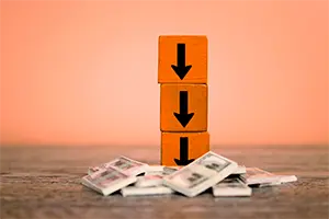 A stack of three orange wooden blocks with downward facing arrows printed on each block. The stack is positioned on a wooden surface with a pile of crumpled banknotes at its base.